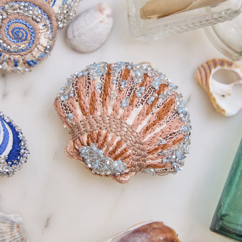 Craftspring’s Scallop Shell Ornament — made of satin with intricate beaded and embroidered details — displayed among seashells and glass bottles, creating a festive coastal holiday scene, ethically crafted by artisans.