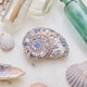 Craftspring’s Moon Snail Shell Ornament — made of satin with intricate beaded and embroidered details — displayed among seashells and glass bottles, creating a festive coastal holiday scene, ethically crafted by artisans.