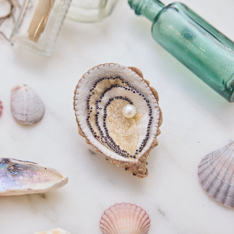 Lifestyle photo of Craftspring’s handmade felt Oyster Christmas ornament, displayed on marble with scattered shells and sea glass bottles—featuring intricate beaded and embroidered details and a shimmering beaded pearl, ethically crafted by artisans using sustainable wool for coastal holiday magic.