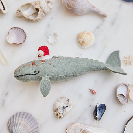 Craftspring’s handmade felt Santa Spyhop Gray Whale Christmas ornament displayed among seashells—featuring intricate embroidered details and a festive red Santa hat, perfect for coastal holiday gifting, ethically crafted by artisans using sustainable wool.