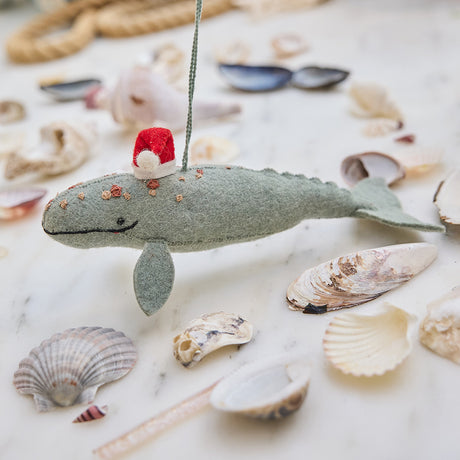 Craftspring’s handmade felt Santa Spyhop Gray Whale Christmas ornament displayed among seashells—featuring intricate embroidered details and a festive red Santa hat, perfect for coastal holiday gifting, ethically crafted by artisans using sustainable wool.