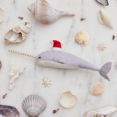 Craftspring’s handmade felt Santa Gray-Blue Narwhal Christmas ornament displayed among seashells—featuring intricate beaded and embroidered details and a festive red Santa hat, perfect for whimsical coastal holiday gifting, ethically crafted by artisans using sustainable wool.