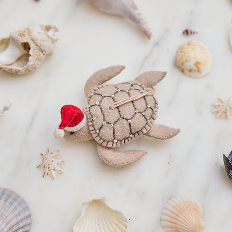 Craftspring’s handmade felt Santa Neutral Sea Turtle Christmas ornament displayed among seashells—featuring intricate beaded and embroidered details and a red Santa hat, perfect for coastal holiday gifting, ethically crafted by artisans using sustainable wool.