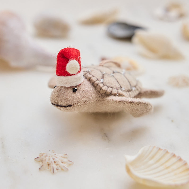 Craftspring’s handmade felt Santa Neutral Sea Turtle Christmas ornament displayed among seashells—featuring intricate beaded and embroidered details and a red Santa hat, perfect for coastal holiday gifting, ethically crafted by artisans using sustainable wool.