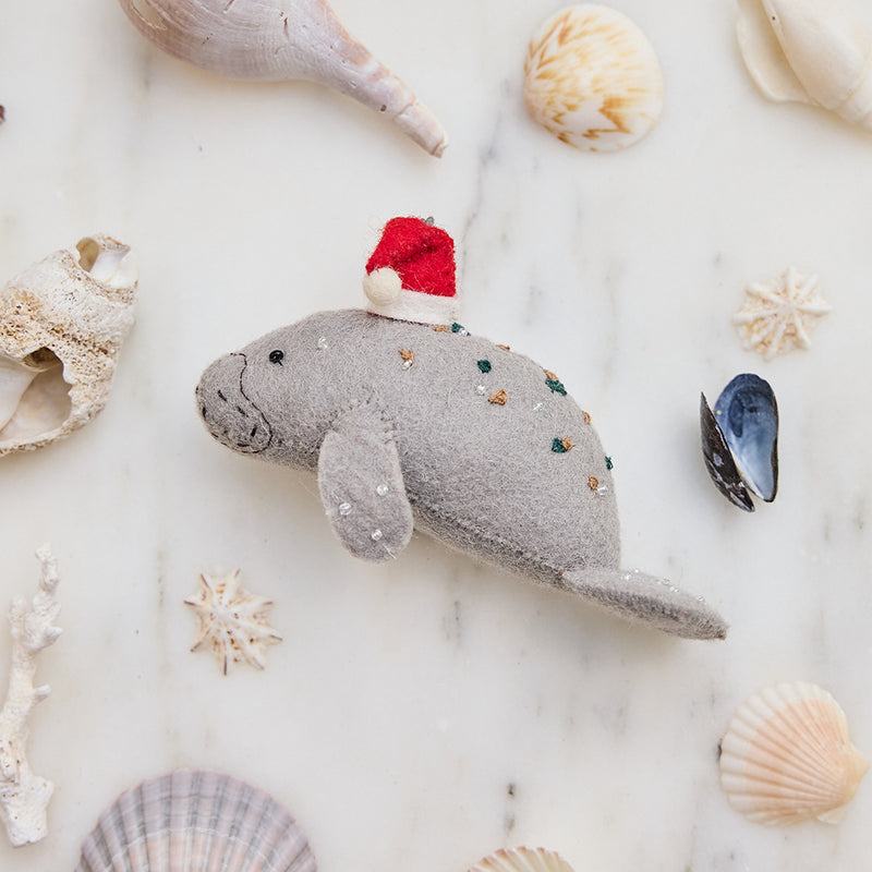 Handmade felt Santa Manatee ornament from Craftspring, featuring a red Santa hat and intricate beaded and embroidered details—displayed among shells for a festive coastal Christmas vibe, ethically crafted by artisans using sustainable wool.