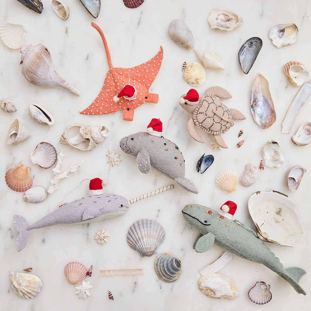 Handmade felt Santa Manatee, Santa Spyhop Gray Whale, Santa Neutral Sea Turtle, Santa Coral Manta, and Santa Gray Blue Narwhal ornaments from Craftspring—each featuring festive Santa hats and intricate beaded and embroidered details, displayed among shells for a joyful coastal Christmas scene, ethically crafted by artisans using sustainable wool.