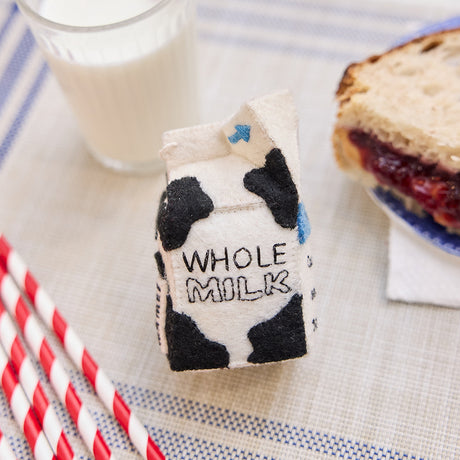 Lifestyle photo of Craftspring’s handmade felt School Milk Carton ornament, featuring embroidered and appliqué details—displayed with a PB&J sandwich and a glass of milk, perfect for nostalgic holiday gifting and lovingly crafted by artisans using sustainable wool.