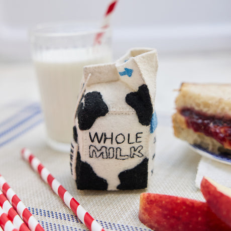 Lifestyle photo of Craftspring’s handmade felt School Milk Carton ornament, featuring embroidered and appliqué details—displayed with a PB&J sandwich, apple slices, and a glass of milk, perfect for nostalgic holiday gifting and lovingly crafted by artisans using sustainable wool.