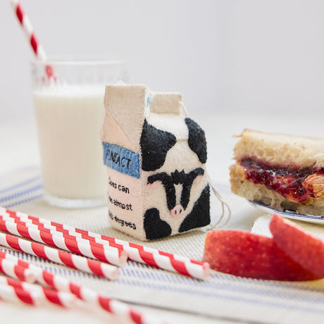 Lifestyle photo of Craftspring’s handmade felt School Milk Carton ornament, featuring embroidered and appliqué details—displayed with a PB&J sandwich, apple slices, and a glass of milk, perfect for nostalgic holiday gifting and lovingly crafted by artisans using sustainable wool.