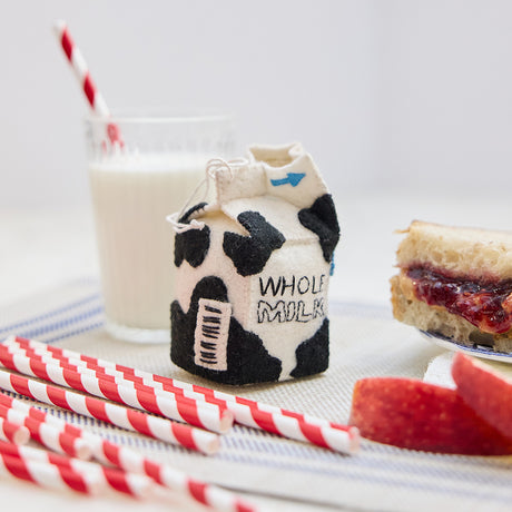 Lifestyle photo of Craftspring’s handmade felt School Milk Carton ornament, featuring embroidered and appliqué details—displayed with a PB&J sandwich, apple slices, and a glass of milk, perfect for nostalgic holiday gifting and lovingly crafted by artisans using sustainable wool.