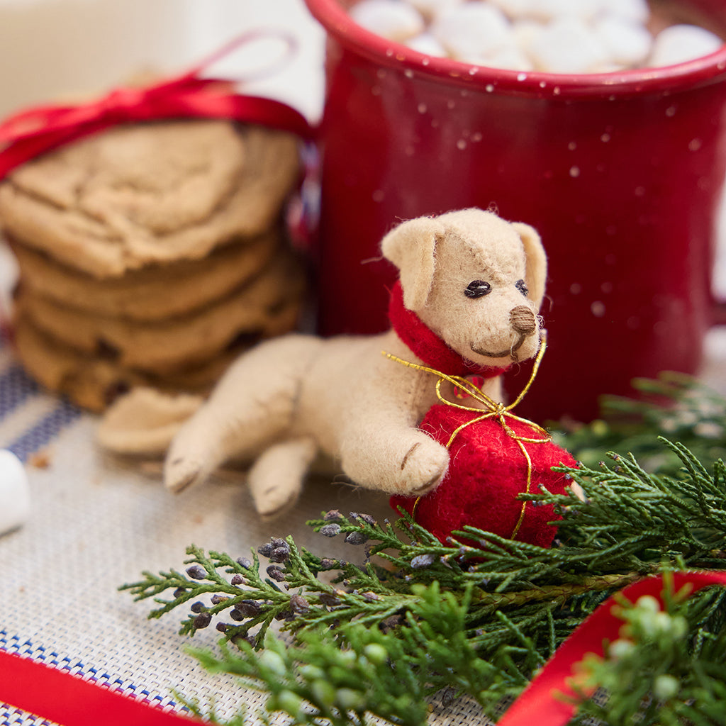 Handmade felt Dog with Gift ornament from Craftspring, featuring a pup holding a red present between its paws—displayed with a mug of hot cocoa and cookies, perfect for festive dog lovers and cozy holiday gifting, ethically crafted by artisans using sustainable wool.