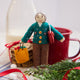 Handmade felt Grandma ornament from Craftspring, wearing a cozy beaded and embroidered holiday sweater, holding a designer purse and a shopping bag full of presents—displayed with milk, cookies, and evergreen branches, evoking warm holiday memories and timeless festive cheer, ethically crafted by artisans using sustainable wool.