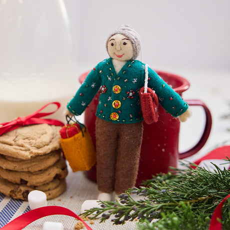 Handmade felt Grandma ornament from Craftspring, wearing a cozy beaded and embroidered holiday sweater, holding a designer purse and a shopping bag full of presents—displayed with milk, cookies, and evergreen branches, evoking warm holiday memories and timeless festive cheer, ethically crafted by artisans using sustainable wool.