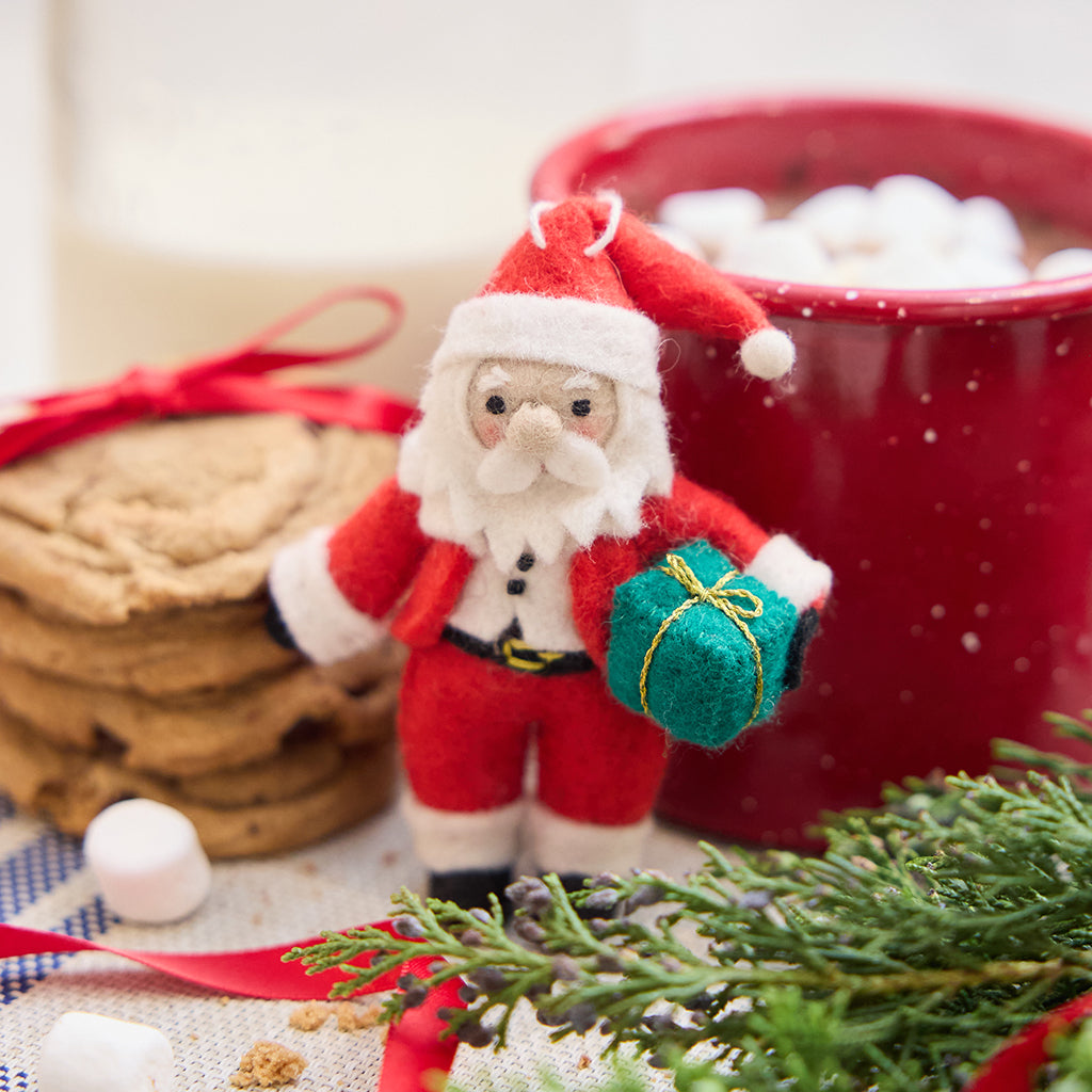 Handmade felt Sweet Little Santa ornament from Craftspring, featuring an adorable Santa with rosy cheeks and a cute round nose holding a gift—displayed with hot cocoa, cookies, and evergreen branches, perfect for cozy Christmas moments and festive gifting, ethically crafted by artisans using sustainable wool.