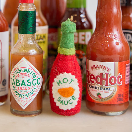 Handmade felt Hot Sauce ornament from Craftspring, featuring bold embroidered details—displayed among real hot sauce bottles, perfect for spicy food lovers and unique holiday gifting, sustainably handmade by artisans using wool.