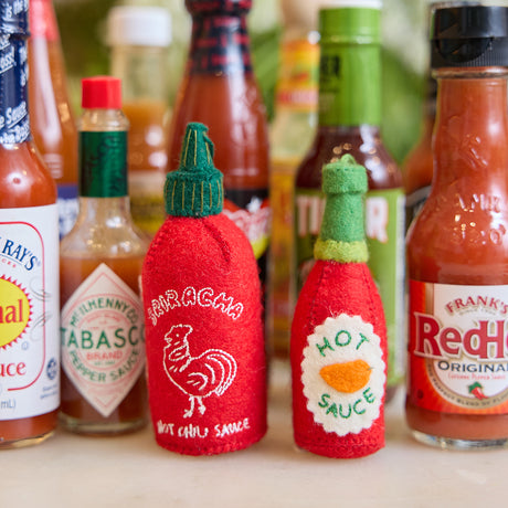 Handmade felt Hot Sauce and Sriracha ornaments from Craftspring, featuring vibrant embroidered details—displayed among hot sauce bottles, perfect for spice lovers and festive holiday gifting, sustainably handmade by artisans using wool.