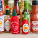 Handmade felt Hot Sauce and Sriracha ornaments from Craftspring, featuring vibrant embroidered details—displayed among hot sauce bottles, perfect for spice lovers and festive holiday gifting, sustainably handmade by artisans using wool.