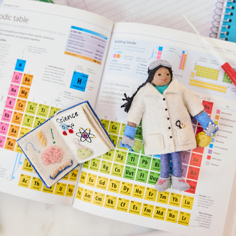 Craftspring’s handmade felt STEAM Book and Young Scientist Christmas ornaments displayed together on a science textbook—featuring intricate beaded and embroidered details that celebrate curiosity and learning, ethically crafted by artisans using sustainable wool.