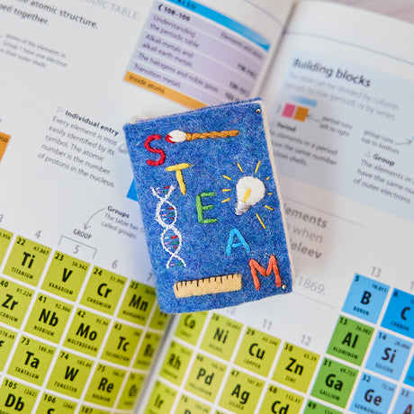 Handmade felt STEAM Book Christmas ornament from Craftspring, featuring an embroidered DNA strand, paintbrush, light bulb, and ruler on the cover—displayed atop a science textbook in a cozy holiday scene, perfect for gifting to curious minds and celebrating creativity and learning with heirloom-quality decor, ethically crafted by artisans using sustainable wool.