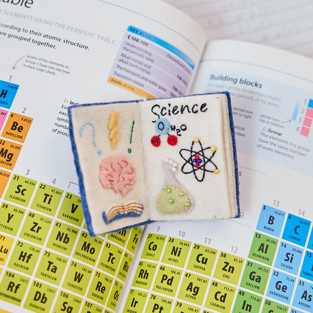 Inside view of Craftspring’s handmade felt STEAM Book Christmas ornament, featuring a “Science” page with intricate beaded and embroidered details—displayed on a science textbook to highlight the ornament’s imaginative design and craftsmanship, ethically crafted by artisans using sustainable wool.