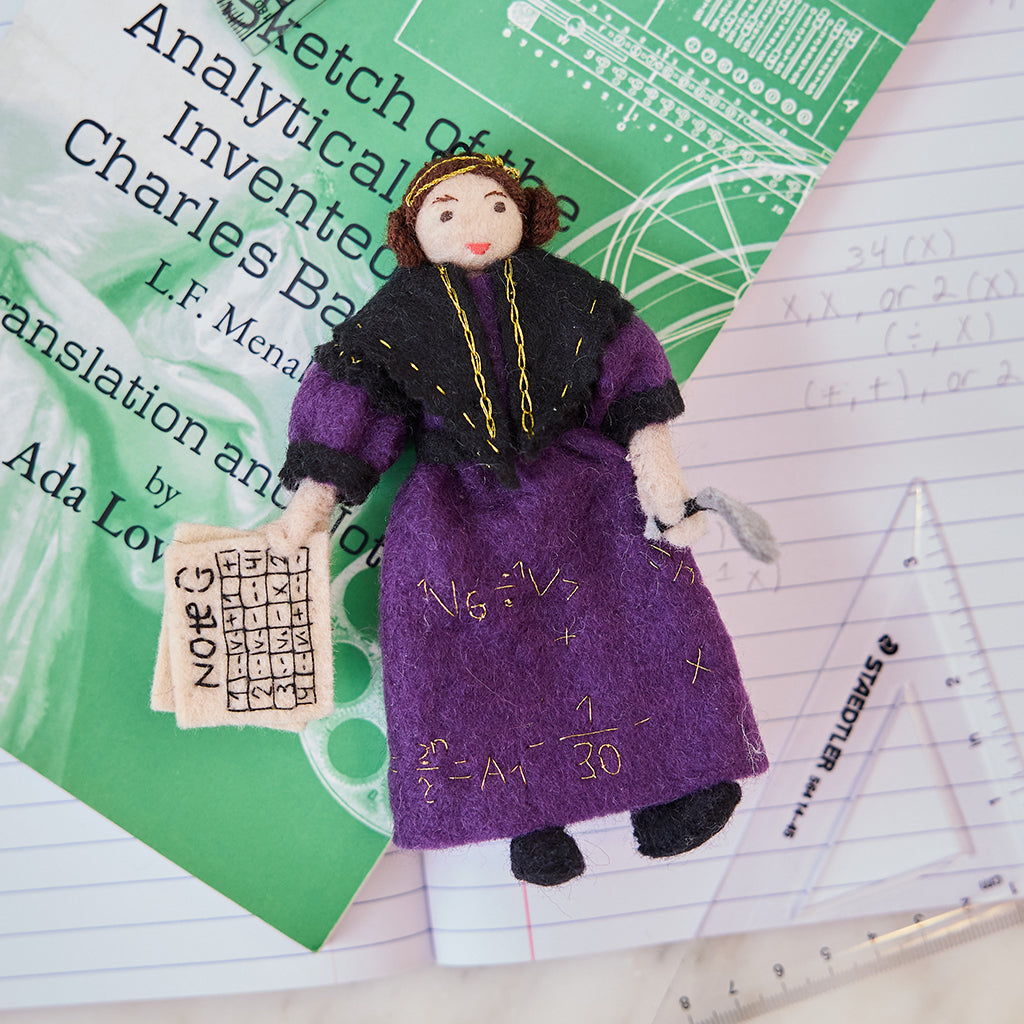 Craftspring’s handmade felt Ada Lovelace Christmas ornament displayed on a book—featuring intricate beaded and embroidered details, including Ada holding Note G and gold embroidered equations on her dress, perfect for thoughtful holiday gifting, ethically crafted by artisans using sustainable wool.