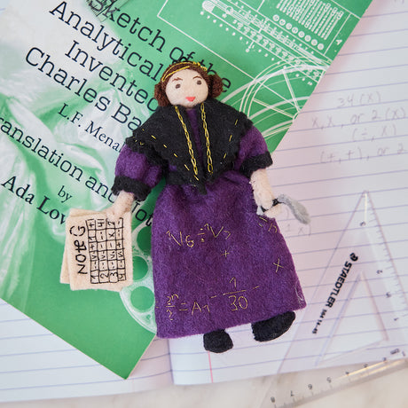 Craftspring’s handmade felt Ada Lovelace Christmas ornament displayed on a book—featuring intricate beaded and embroidered details, including Ada holding Note G and gold embroidered equations on her dress, perfect for thoughtful holiday gifting, ethically crafted by artisans using sustainable wool.