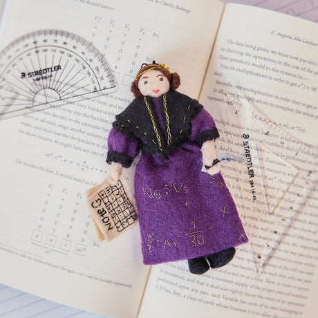 Craftspring’s handmade felt Ada Lovelace Christmas ornament displayed on an open science book—featuring intricate beaded and embroidered details including Ada holding Note G and gold embroidered equations on her dress, perfect for thoughtful holiday gifting, ethically crafted by artisans using sustainable wool.
