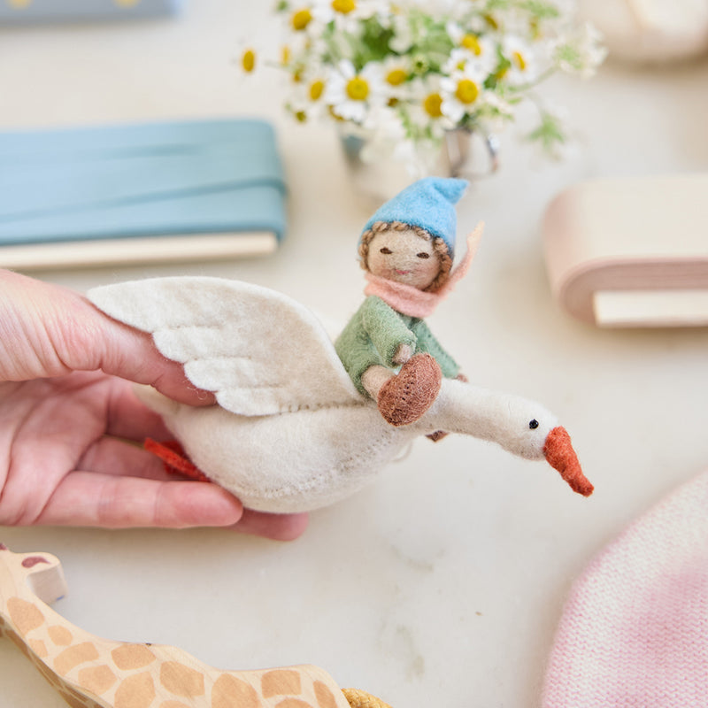Handmade felt Cloud Dancer Dream Goose Ornament – Light from Craftspring held in hand, surrounded by a baby hat, toy, and ribbons—featuring a detailed baby with light skin and blonde hair riding a white goose, perfect for nursery decor and heartfelt baby shower gifting, ethically crafted using sustainable wool.