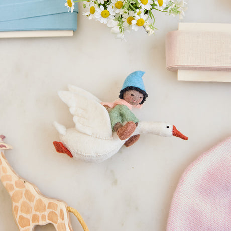 Handmade felt Cloud Dancer Dream Goose ornament – Brown – from Craftspring, displayed on marble with a baby hat, toys, and ribbons—featuring a baby with brown skin and black hair riding a white goose, perfect as a charming nursery decoration and thoughtful keepsake, ethically crafted by artisans using sustainable wool.