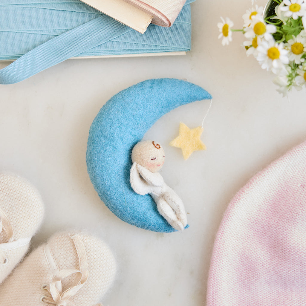 Handmade felt Moon Dreamer Baby Ornament – Light from Craftspring, featuring a sleeping baby with light skin on a crescent moon with a dangling star—displayed on marble surrounded by baby booties, a baby hat, flowers, and ribbons, perfect for nursery decor and meaningful baby shower gifting, ethically crafted using sustainable wool.