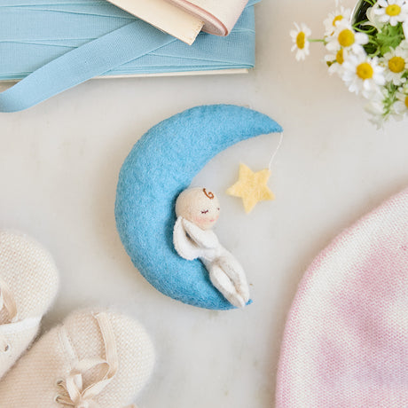 Handmade felt Moon Dreamer Baby Ornament – Light from Craftspring, featuring a sleeping baby with light skin on a crescent moon with a dangling star—displayed on marble surrounded by baby booties, a baby hat, flowers, and ribbons, perfect for nursery decor and meaningful baby shower gifting, ethically crafted using sustainable wool.