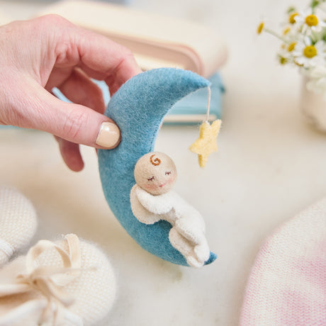 Handmade felt Moon Dreamer Baby Ornament – Light from Craftspring, held in hand and surrounded by baby booties, a baby hat, and flowers—featuring a sleeping baby with light skin on a crescent moon with a dangling star, perfect for nursery decor and heartfelt baby shower gifting, ethically crafted using sustainable wool.