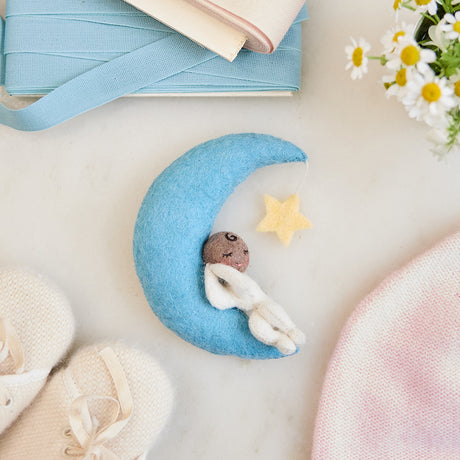 Handmade felt Moon Dreamer Baby Ornament – Brown from Craftspring, featuring a sleeping baby with brown skin on a crescent moon with a dangling star—displayed on marble surrounded by baby booties, a baby hat, flowers, and ribbons, perfect for nursery decor and meaningful baby shower gifting, ethically crafted using sustainable wool.