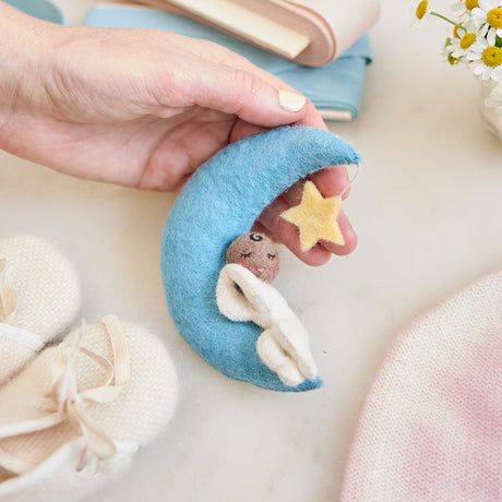 Handmade felt Moon Dreamer Baby Ornament – Brown from Craftspring, featuring a sleeping baby with brown skin on a crescent moon with a dangling star—held in hand and surrounded by baby booties, a baby hat, and flowers, perfect for nursery decor and heartfelt baby shower gifting, ethically crafted by artisans using sustainable wool.