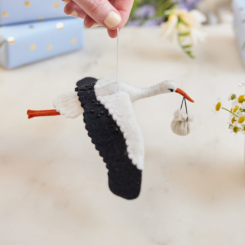 Handmade felt New Baby Stork ornament from Craftspring, featuring a stork carrying a bundle with delicate beaded and embroidered details—displayed hanging surrounded by flowers and presents, perfect for nursery decor and thoughtful baby shower gifting, ethically crafted by artisans using sustainable wool.
