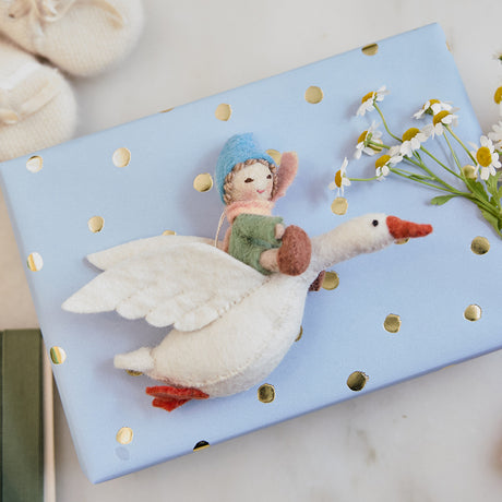 Handmade felt Cloud Dancer Dream Goose Ornament – Light from Craftspring, displayed on a wrapped gift with baby booties nearby—featuring a detailed baby with light skin and blonde hair riding a flying white goose, perfect for nursery decor and thoughtful baby shower gifting, ethically crafted using sustainable wool.