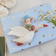 Handmade felt Cloud Dancer Dream Goose Ornament – Light from Craftspring, displayed on a wrapped gift with baby booties nearby—featuring a detailed baby with light skin and blonde hair riding a flying white goose, perfect for nursery decor and thoughtful baby shower gifting, ethically crafted using sustainable wool.