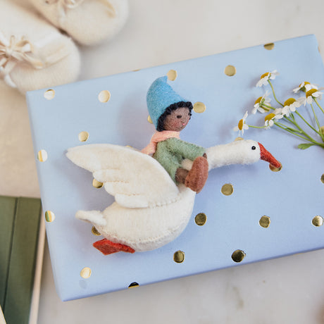 Handmade felt Cloud Dancer Dream Goose ornament – Brown – from Craftspring, displayed on a wrapped gift with baby booties nearby—featuring a baby with brown skin and black hair riding a white goose, perfect for nursery decor and heartfelt baby shower gifting, ethically crafted using sustainable wool.