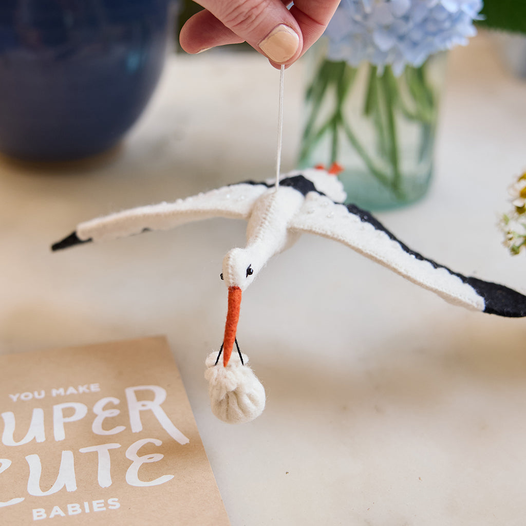 Handmade felt New Baby Stork ornament from Craftspring, featuring a stork carrying a bundle with delicate beaded and embroidered details—displayed hanging surrounded by flowers and a card, perfect for nursery decor and thoughtful baby shower gifting, ethically crafted by artisans using sustainable wool.
