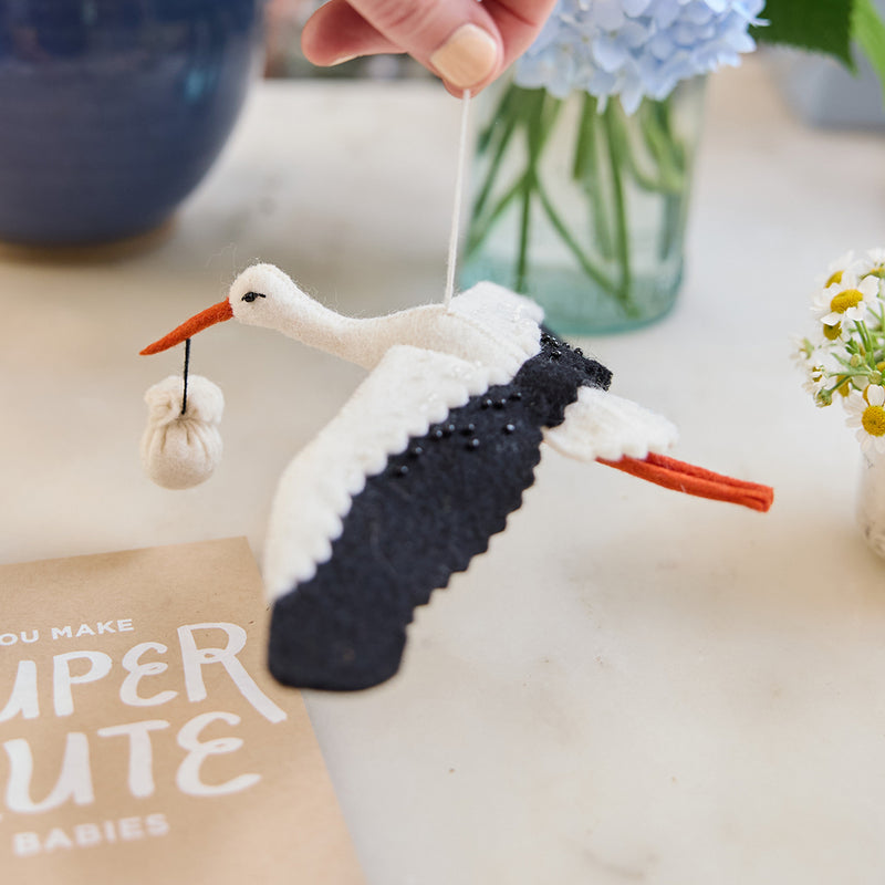 Handmade felt New Baby Stork ornament from Craftspring, featuring a stork carrying a bundle with delicate beaded and embroidered details—displayed hanging surrounded by flowers and a card, perfect for nursery decor and thoughtful baby shower gifting, ethically crafted by artisans using sustainable wool.