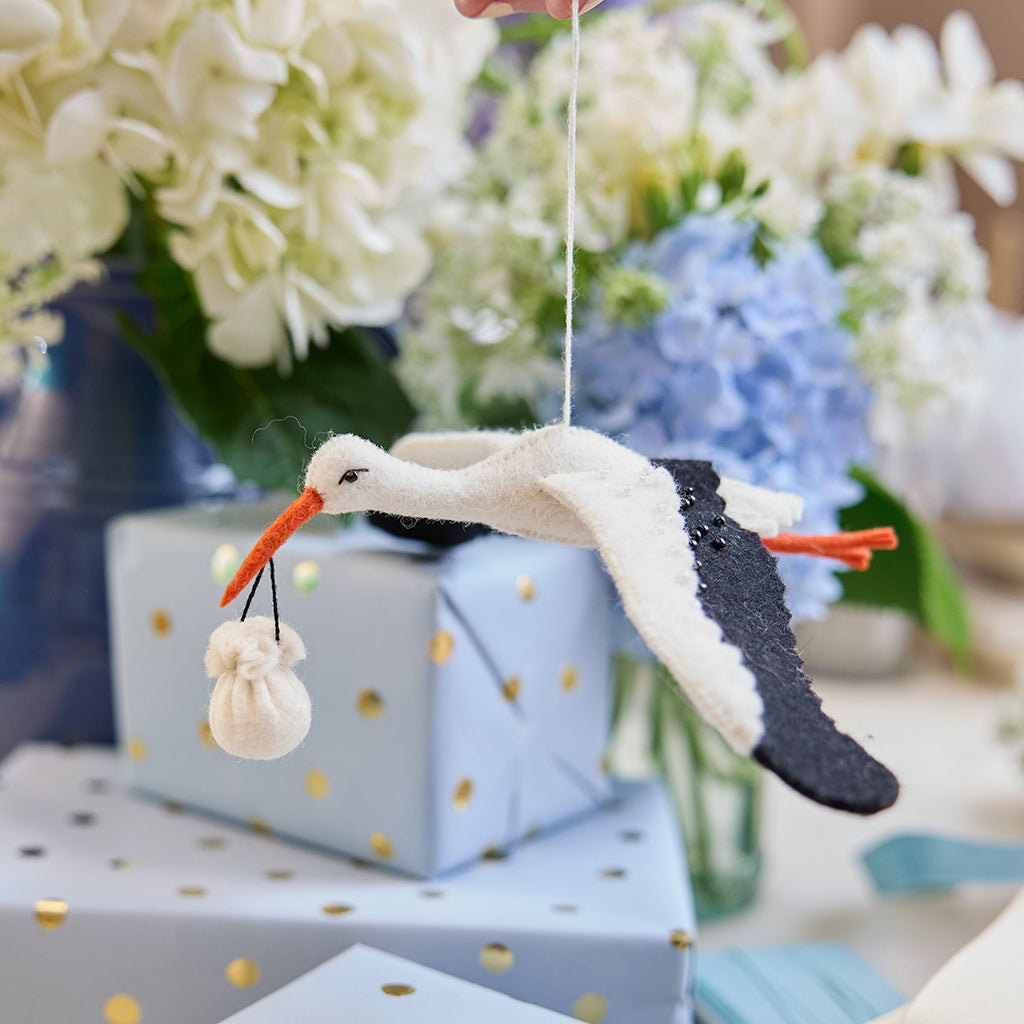 Handmade felt New Baby Stork ornament from Craftspring, featuring a stork carrying a bundle with delicate beaded and embroidered details—displayed hanging in front of flowers and presents, perfect for nursery decor and thoughtful baby shower gifting, ethically crafted by artisans using sustainable wool.