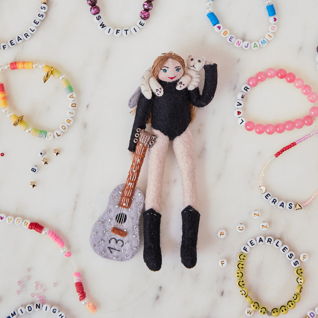 Craftspring’s handmade felt Taylor Swift with Cat Christmas ornament displayed surrounded by friendship bracelets—featuring Taylor holding a guitar with the number 13 and a cat around her shoulders, perfect for Swiftie holiday gifting, ethically crafted by artisans using sustainable wool.
