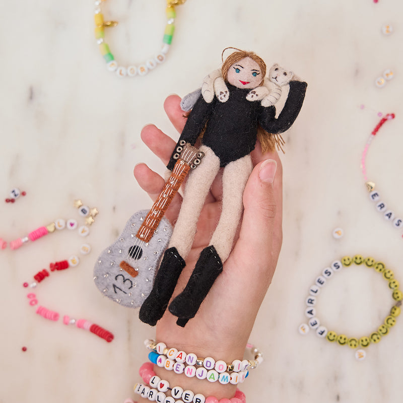 Craftspring’s handmade felt Taylor Swift with Cat Christmas ornament displayed surrounded by friendship bracelets—featuring Taylor holding a guitar with the number 13 and a cat around her shoulders, perfect for Swiftie holiday gifting, ethically crafted by artisans using sustainable wool.