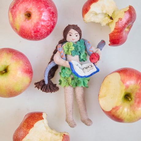 Craftspring’s handmade felt Eve Christmas ornament displayed surrounded by apples and apple cores—featuring intricate details of Eve holding a book of knowledge and an apple, wrapped in leaves with a serpent around her shoulders, perfect for thoughtful holiday gifting, ethically crafted by artisans using sustainable wool.