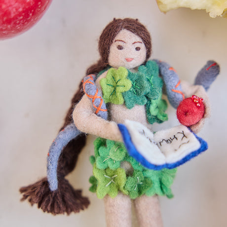 Craftspring’s handmade felt Eve Christmas ornament displayed surrounded by apples and apple cores—featuring intricate details of Eve holding a book of knowledge and an apple, wrapped in leaves with a serpent around her shoulders, perfect for thoughtful holiday gifting, ethically crafted by artisans using sustainable wool.