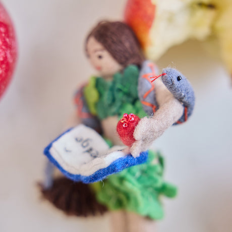 Craftspring’s handmade felt Eve Christmas ornament displayed surrounded by apples and apple cores—featuring intricate details of Eve holding a book of knowledge and an apple, wrapped in leaves with a serpent around her shoulders, perfect for thoughtful holiday gifting, ethically crafted by artisans using sustainable wool.