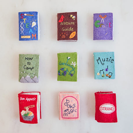Craftspring’s collection of nine handmade felt book Christmas ornaments—STEAM, Nature Guide, Craft, How to Camp, Gardening, Music, Bon Appetit, Books Are Magic, and Strand Bookstore Book—each featuring intricate beaded and embroidered details with pages that open to reveal detailed interiors, displayed together to celebrate creativity and the joy of reading, ethically crafted by artisans using sustainable wool.