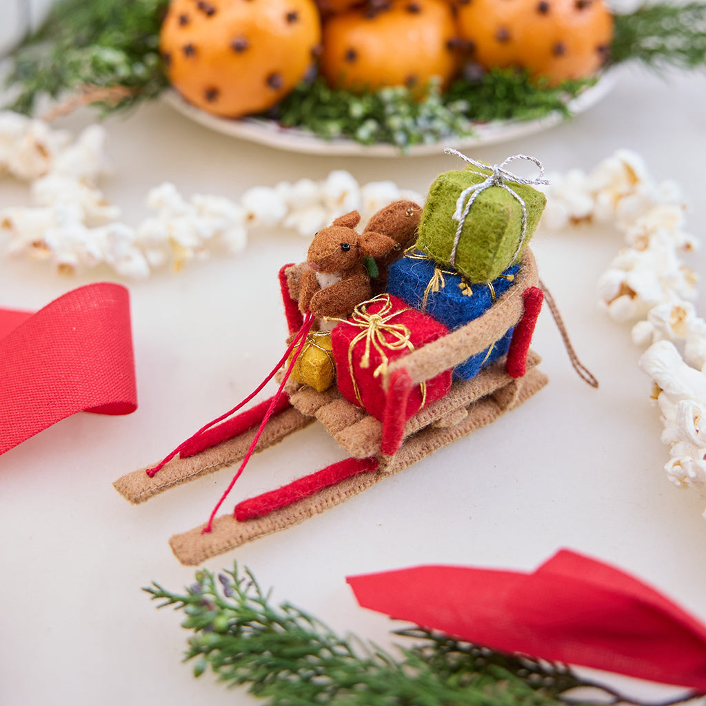 Handmade felt Squirrel with Presents on Sled ornament from Craftspring, featuring a squirrel riding a vintage sled filled with presents—displayed among evergreens, ribbons, and a popcorn garland, perfect for festive Christmas gifting and charming holiday decor, ethically crafted by artisans using sustainable wool.