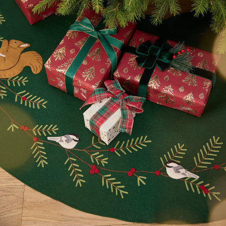 Craftspring’s Woodland Tree Skirt displayed under a Christmas tree with presents — close-up of the embroidered and appliqué chickadees and squirrel among pine branches and red berries, highlighting festive holiday details, ethically crafted by artisans using sustainable wool.