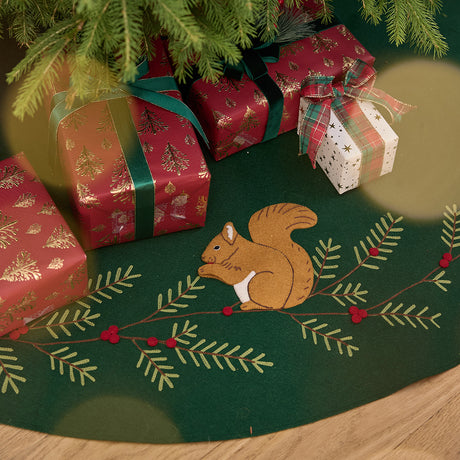 Craftspring’s Woodland Tree Skirt displayed under a Christmas tree with presents — close-up of the embroidered and appliqué squirrel nestled among pine branches and red berries, highlighting festive holiday details, ethically crafted by artisans using sustainable wool.